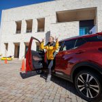 Woman in yellow jacket standing in front of car facing forward with horns up.